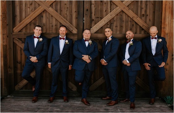 groomsmen in navy suits maroon ties greenery and white flower corsage for november wedding colors 2023 maroon and green