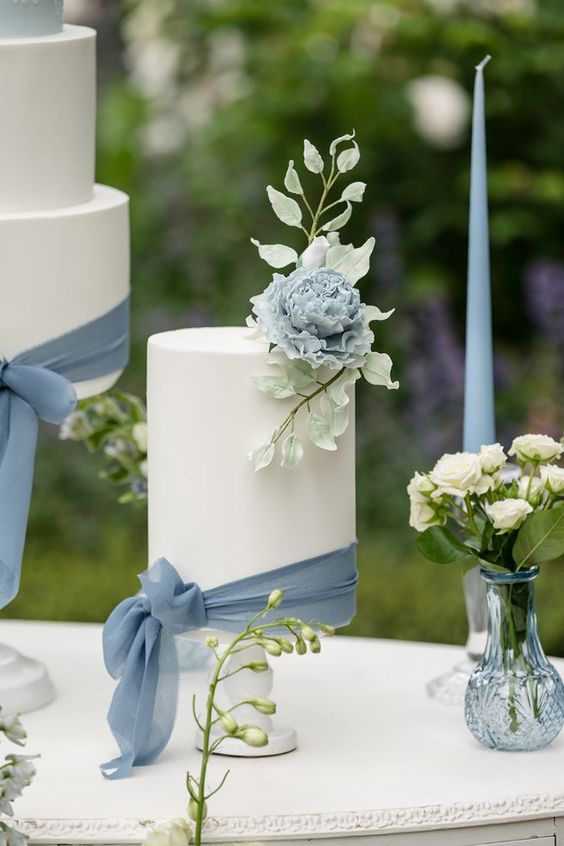 white wedding cake dotted with dusty blue sash and flowers for dusty blue wedding themes for 2023 dusty blue and navy blue