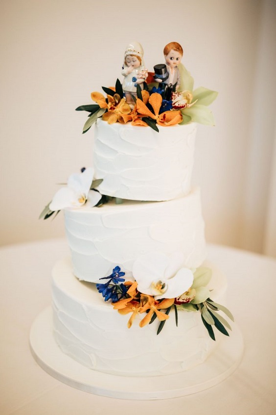 white wedding cake dotted with orange flower and greenery for royal blue wedding colors 2024 royal blue and orange