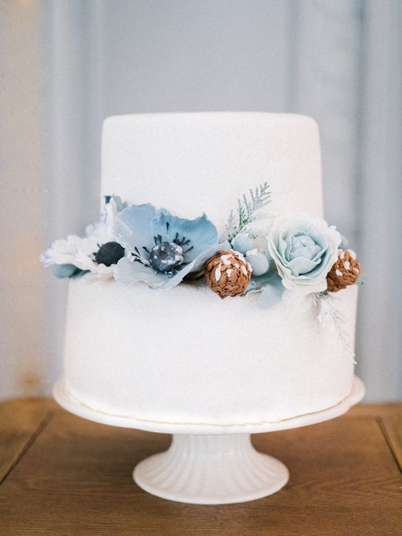 white wedding cake dotted with dusty blue and light blue flowers and pine cones for december wedding colors 2024 shades of blue
