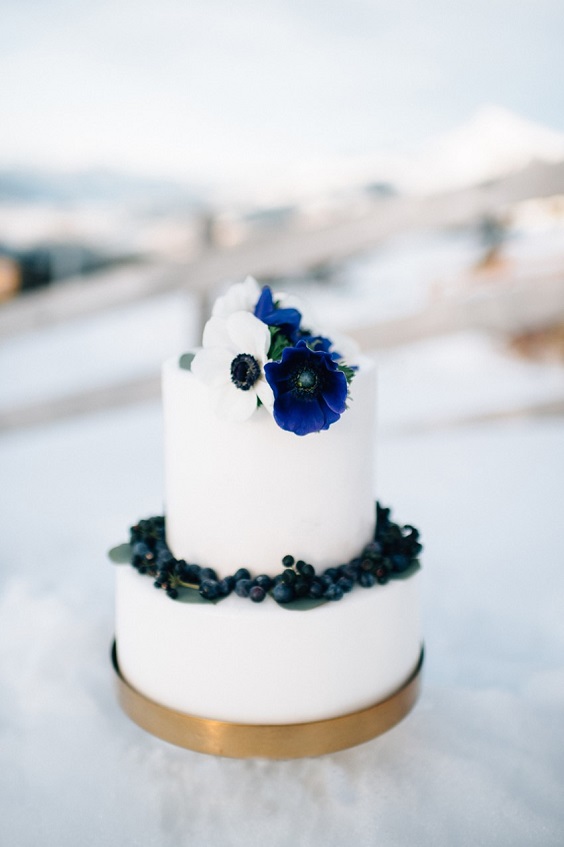 white wedding cake dotted with navy blue flowers and blueberries and gold circle for january wedding colors for 2024 navy blue gold and white